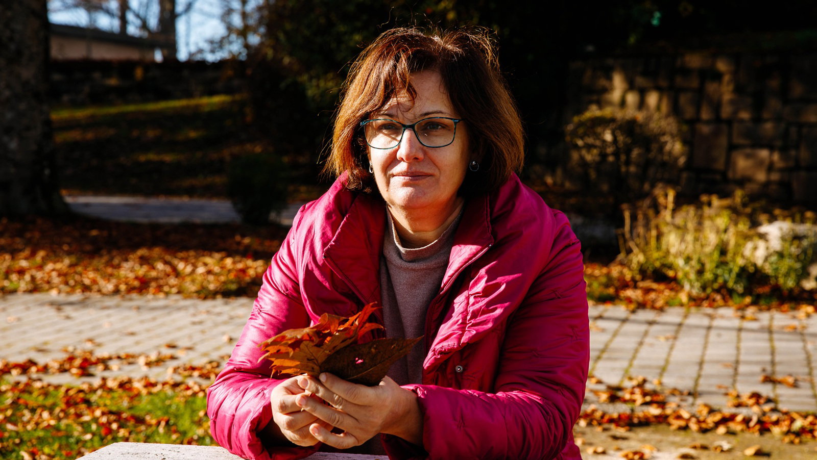 kobieta w wieku 50 plus w okularach siedzi na tle jesiennej alejki z bukietem suchych liści w ręku zamyślona