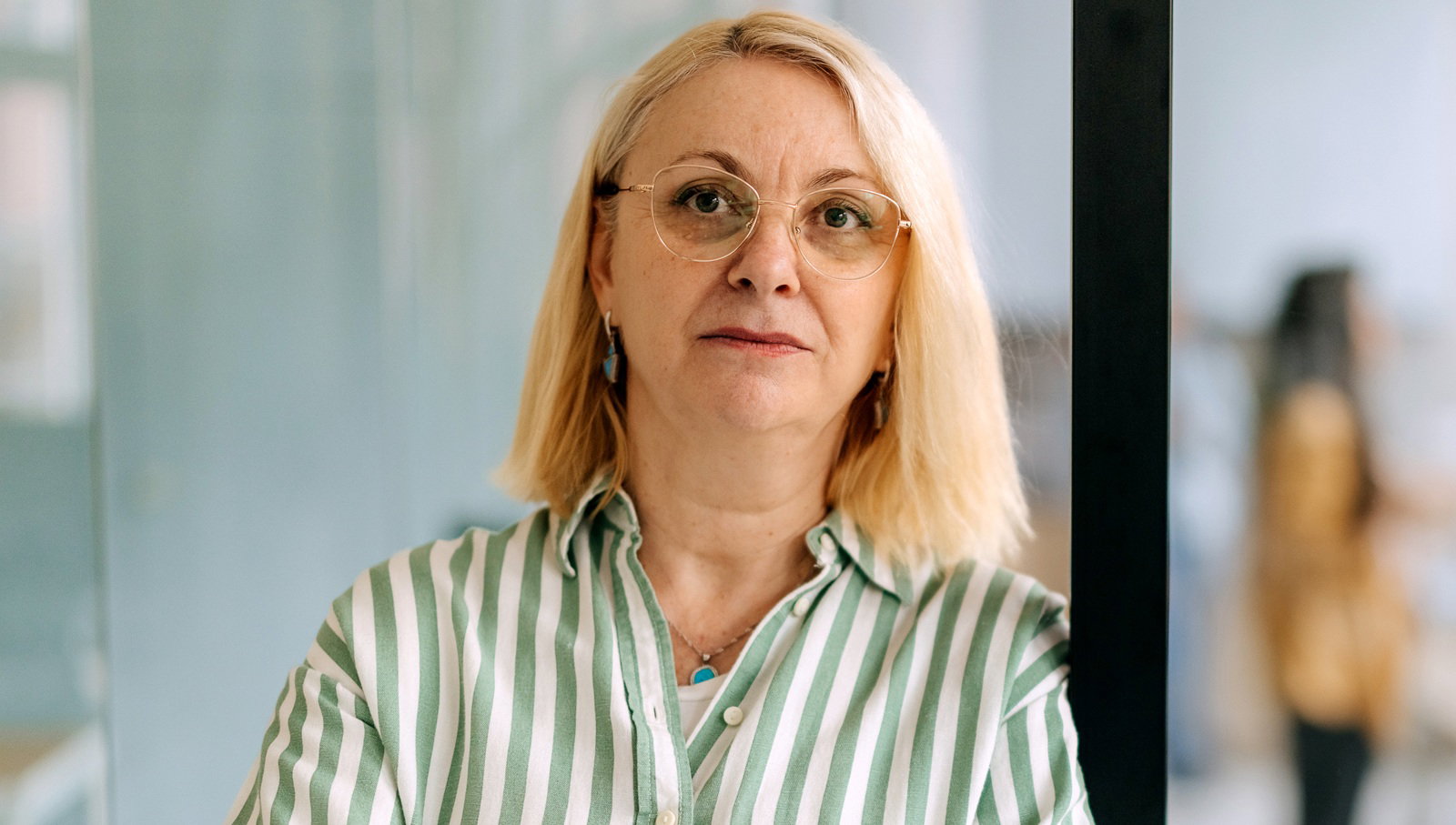 kobieta w wieku 50 plus z pasiastej koszuli i w okularach blondynka stoi na tle szklanej szyby z poważną miną