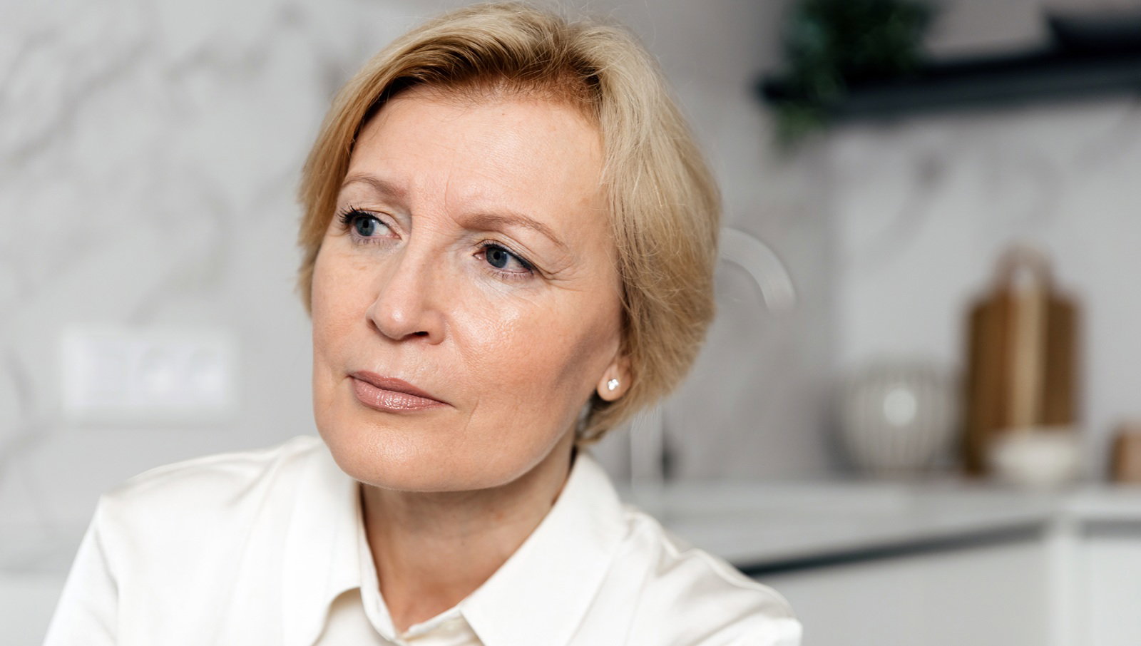 kobieta w wieku 55 plus blondynka z krótkimi włosami elegancka siedzi na tle kuchni lekko zaniepokojona