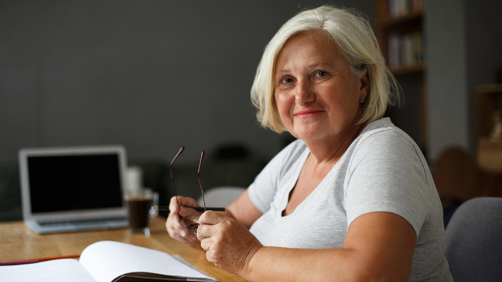 kobieta w wieku 60 plus z blond włosami siedzi przy biurku trzyma w rękach okulary i uśmiecha się zadowolona