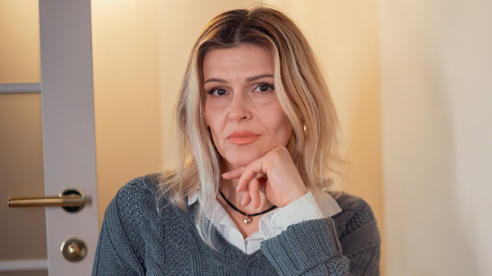 kobieta w wieku 35 plus blondynka w szarym swetrze siedzi przy stole z poważna i surową miną na tle salonu