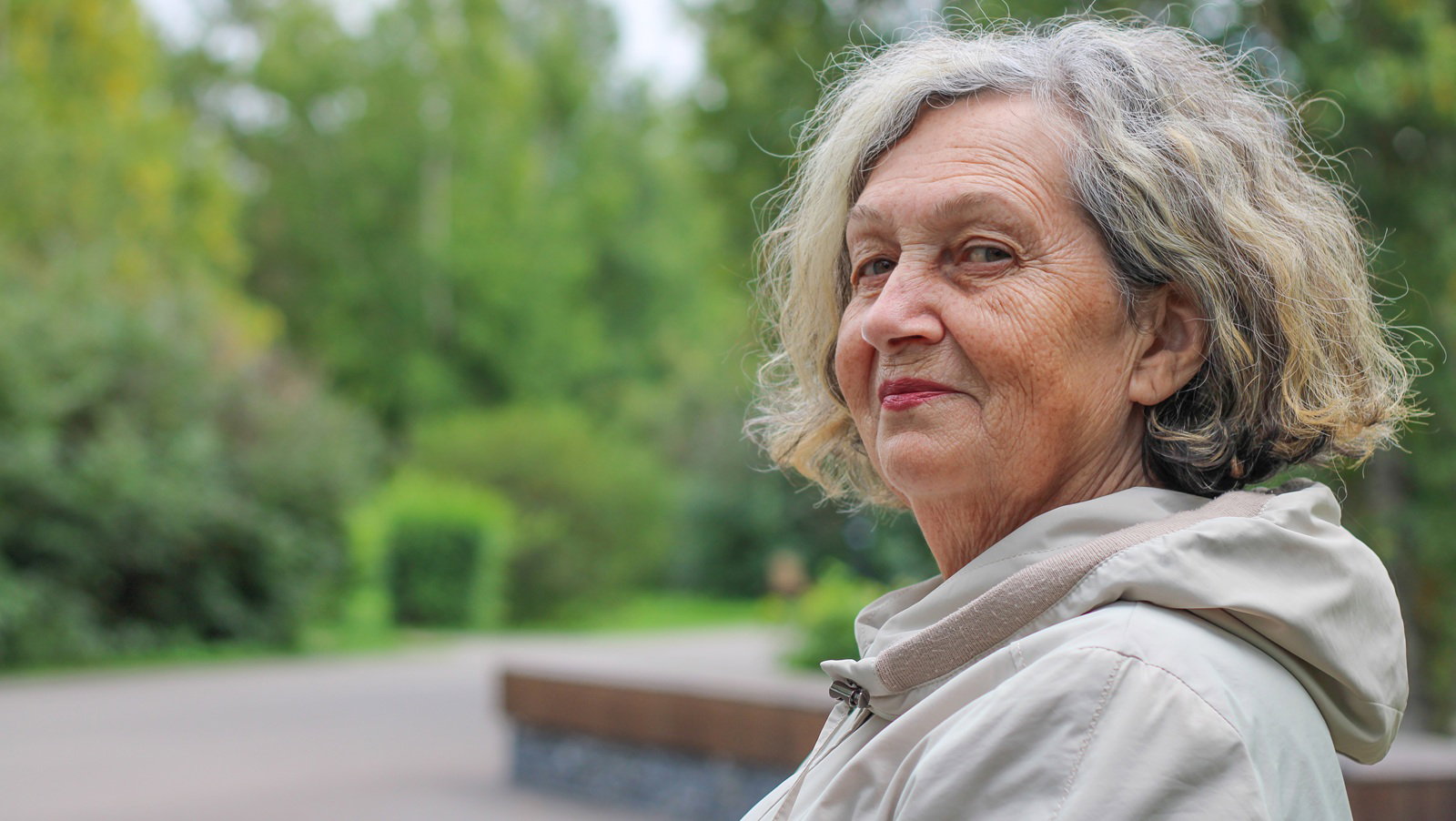 kobieta w wieku 65 plus z siwiejącymi włosami siedzi w jasnej kurtce na tle jesiennego parku i lekko się uśmiecha