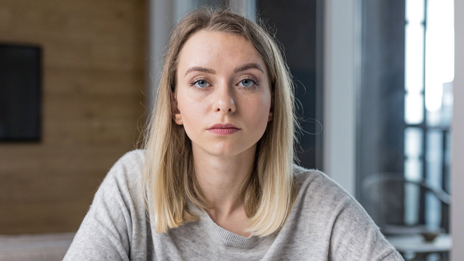 kobieta w wieku 30 plus blondynka w szarym luźnym sweterku siedzi na tle pokoju i okna ze zdegustowaną miną