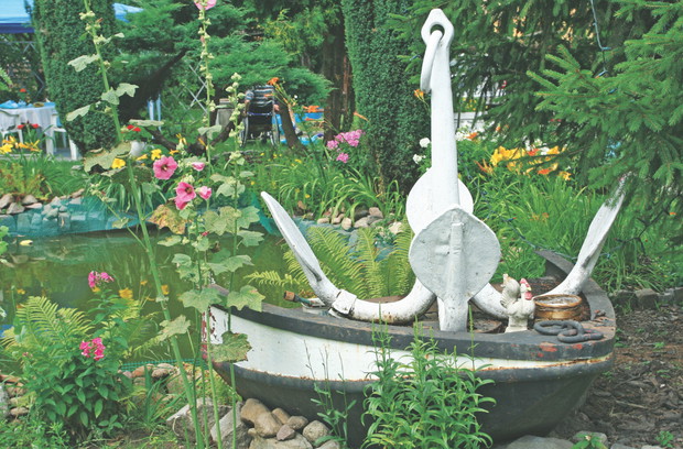 Wokół oczka wodnego rosną paprocie i kwiaty w doniczkach.