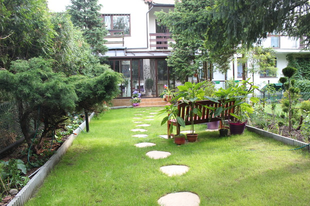 widok z końca ogrodu na dom i ogród zimowy. Z uwagi na dość mało słoneczny układ ogrodu większość lewej strony na tym zdjęciu to rośliny cieniolubne jak kilka odmian funkii, żurawki, żuraweczki i oczywiście różnokolorowe paprocie. Niestety  przekwitł już 