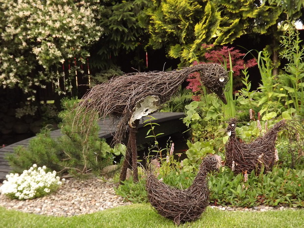 Na tle roślin i wody stoją precyzyjnie wykonane zwierzęta, kura,kogut i bocian. 
