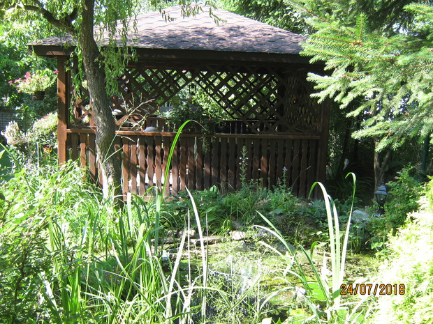 Widok altany od strony południowej , poniżej altany zarys skalniaka okalającego z dwóch stron małe oczko wodne .