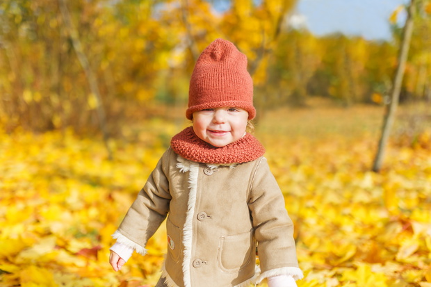 Little smiling girl in knitted cap with fallen autumn leaves is  in city park.