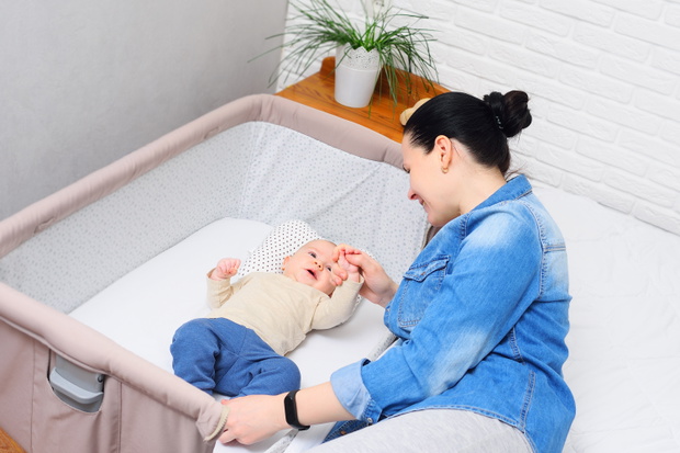 mother plays with a baby who is lying in a side-bed for a newborn or in a modern playpen