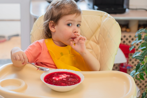 A small child eats vegetable soup with a spoon while sitting on a high chair