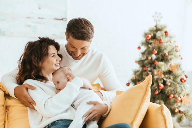 happy man hugging wife sitting on yellow sofa with baby near christmas tree