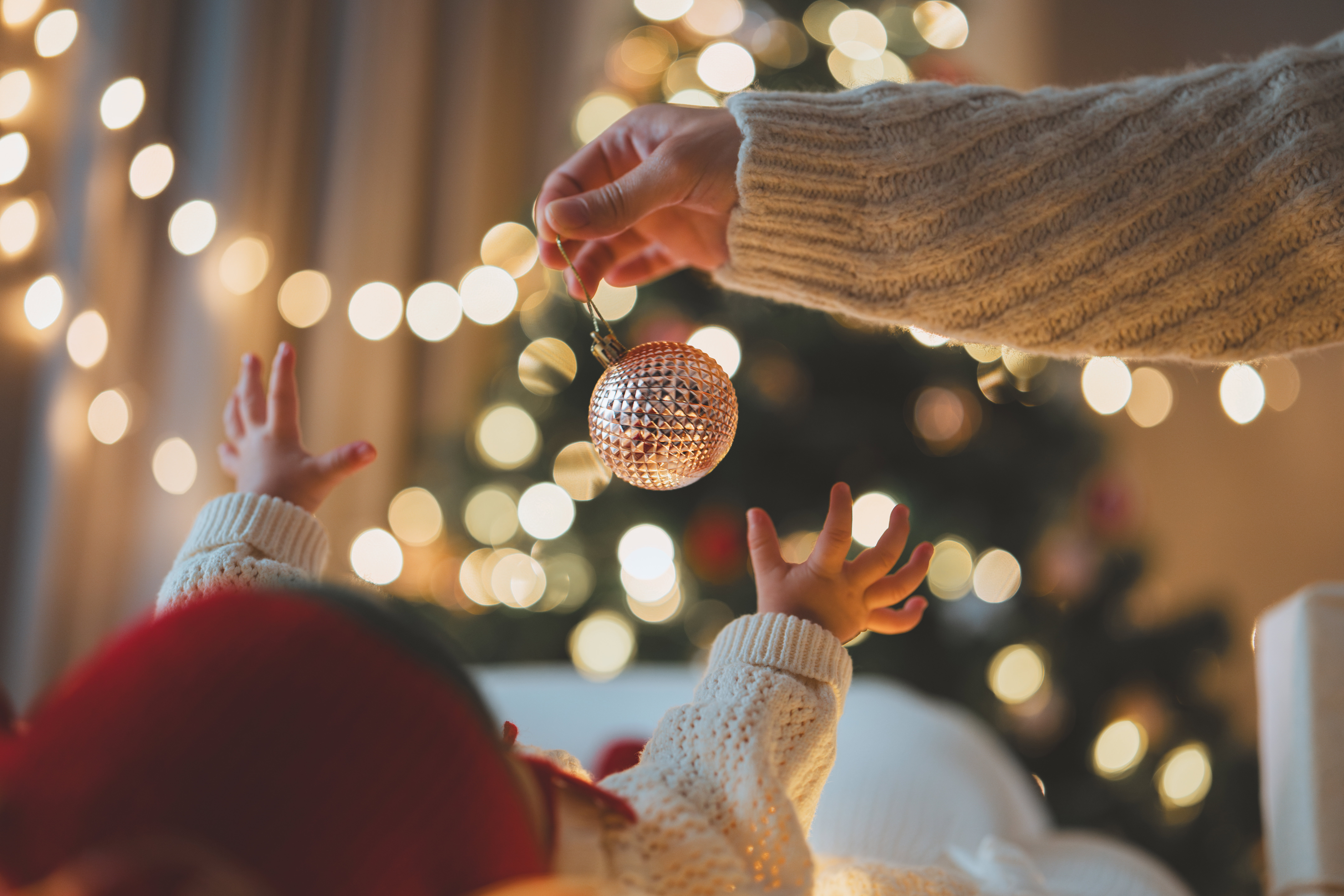 Baby reaching out for a Christmas ornament under glowing tree lights, celebrating first Christmas with parent in warm festive atmosphere filled with love and joy.