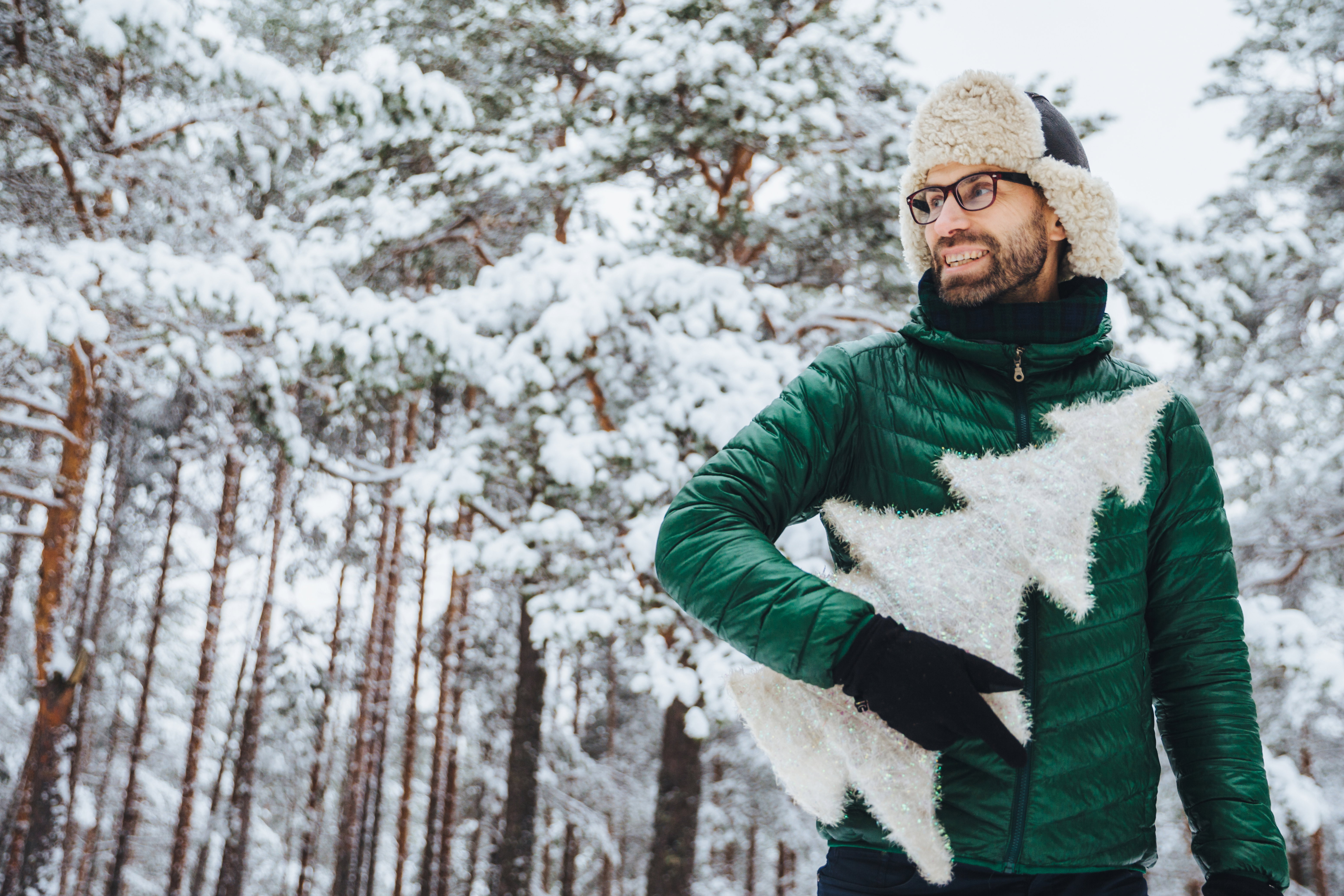 Cheerful pleasant looking man with beard looks thoughtfully aside, holds fir tree, wears spectacles, hat and jacket, poses against winter trees covered with snow. People and lifestyle concept
