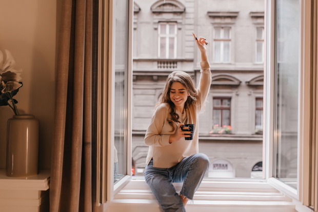 Joyful smiling woman sitting on sill and waving hand. indoor portrait of fashionable young lady with curly hair posing near window with cup of hot bevegare..