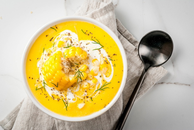 Vegan cuisine. Traditional autumn corn soup. Served with corn cobs, greens, cream. On a white marble table. Copy space top view