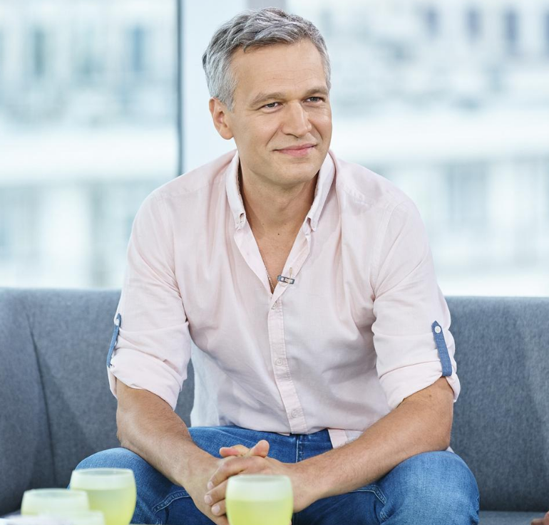 Michał Żebrowski siedzi na szarej kanapie