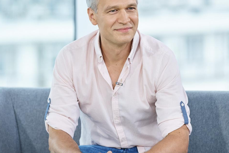 Michał Żebrowski siedzi na szarej kanapie
