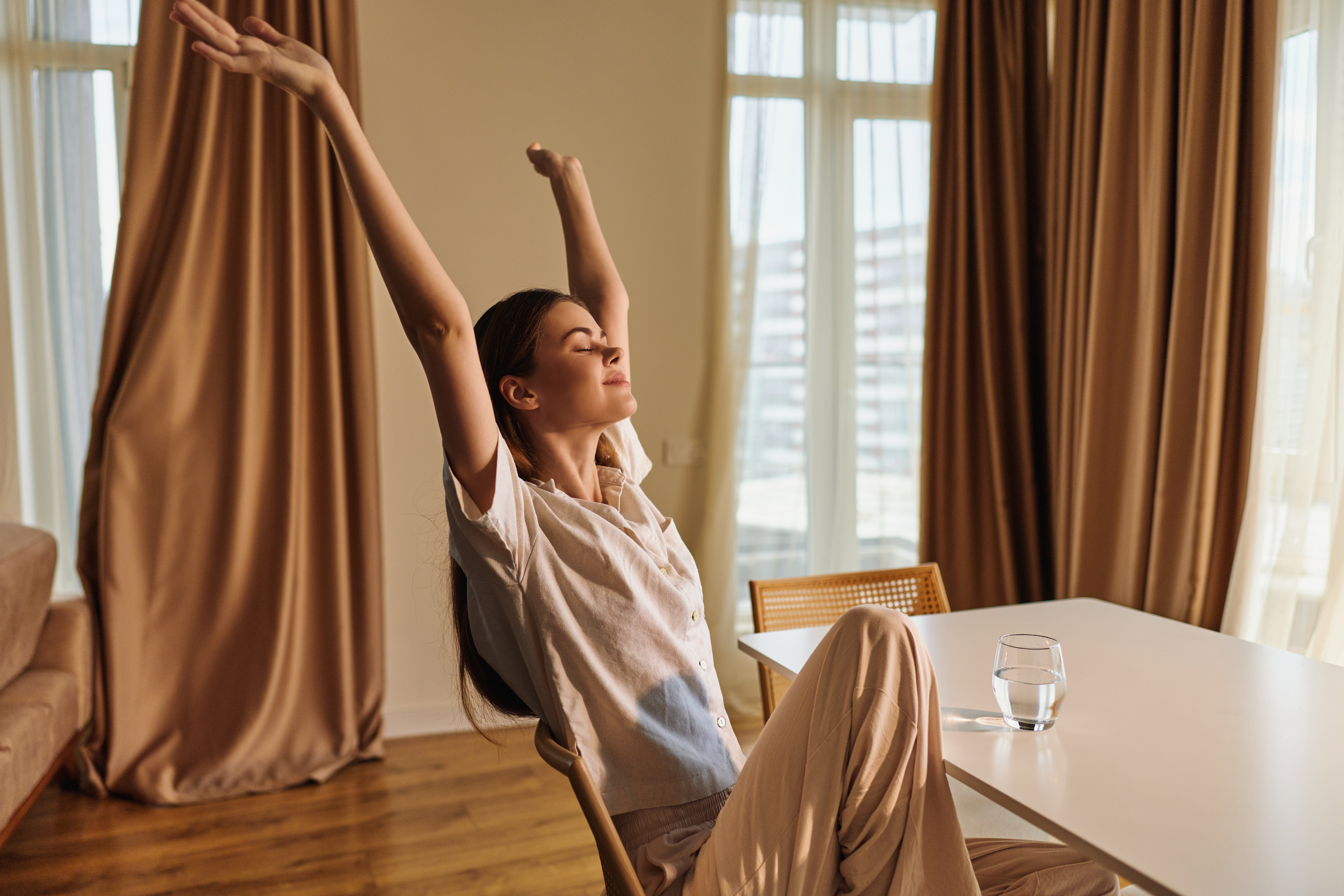 Woman relaxed stretching in morning at home, sitting on chair by table with glass of water, casual clothes and peaceful expression, natural light and cozy interior