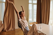 Woman relaxed stretching in morning at home, sitting on chair by table with glass of water, casual clothes and peaceful expression, natural light and cozy interior