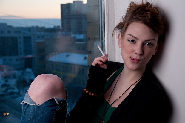 Beautiful young woman smoking on evening window.