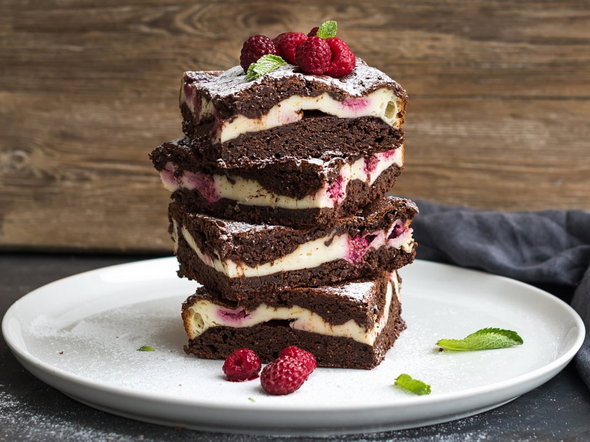 Brownies-cheesecake tower with raspberries on white plate, wooden backdrop, copy space