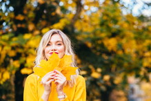 Autumn walk. Woman portrait. Happy girl in yellow hoodie and jean jacket is playing with leaf, looking at camera and smiling while lying on the ground in the park top view