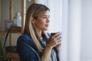 Young woman holding a cup of coffee standing by the window and looking out
