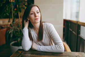 Attractive caucasian dreamful woman in light casual clothes sitting alone near big window in coffee shop, relaxing in restaurant during free time. Young female having rest in cafe. Lifestyle concept.