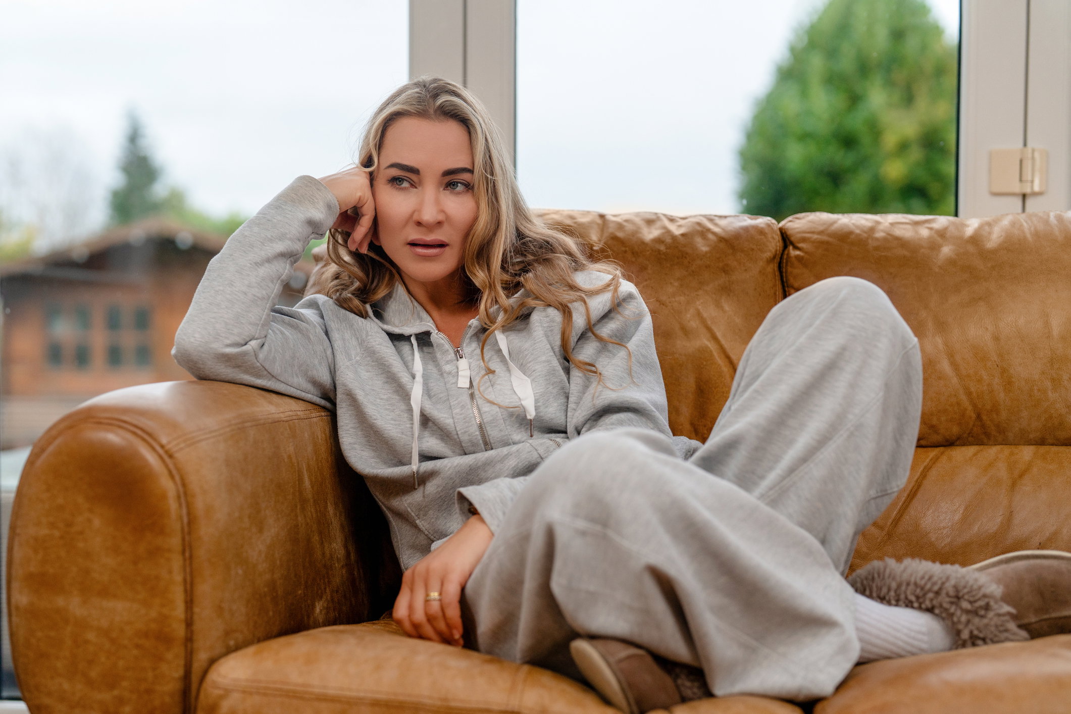 Woman sits on brown couch wearing gray clothing and resting her chin on her hand in living room