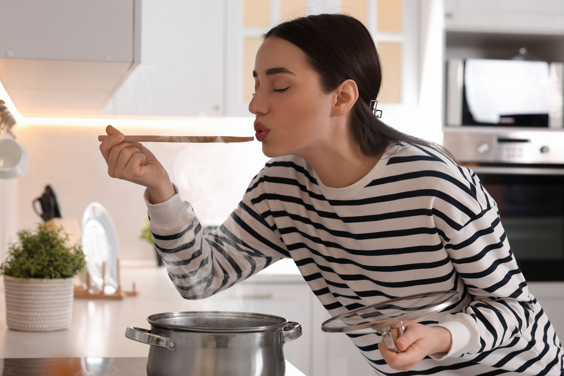 Beautiful woman with wooden spoon tasting soup in kitchen