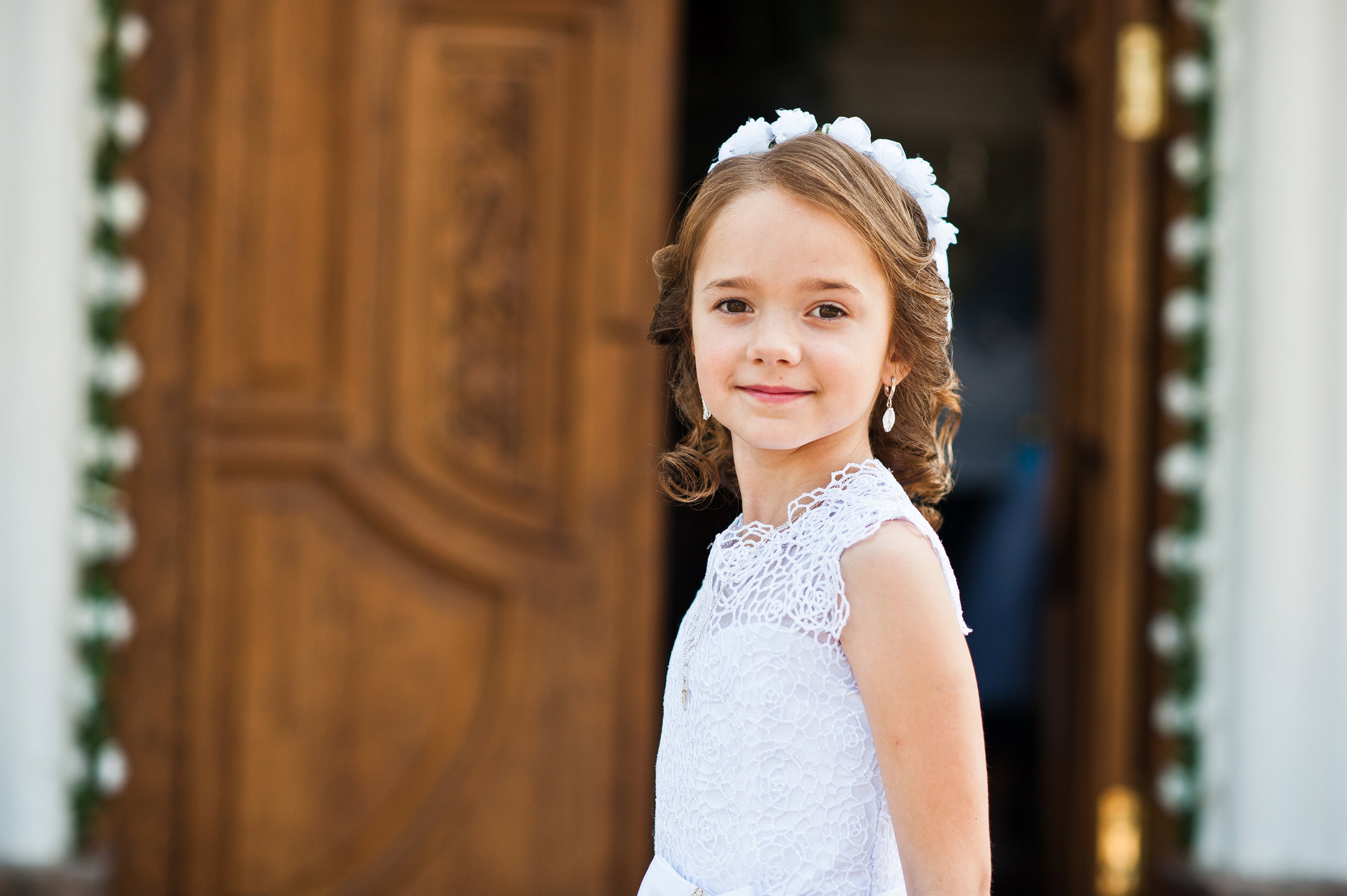 Portrait of cute little girl on white dress and wreath on first holy communion background church gate