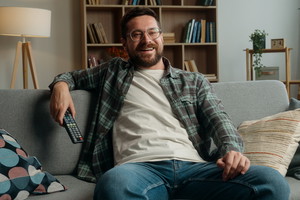 Smiling happy carefree Caucasian man male guy at home couch sofa indoors holiday weekend resting relaxing switch channels with remote control watching TV watch television program film movie sport game