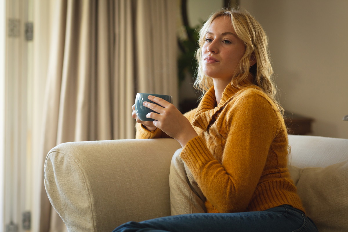 Thoughtful caucasian woman sitting on couch in living room holding coffee cup