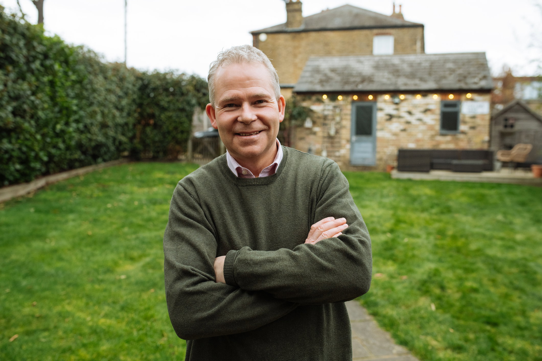 Senior homeowner in UK with arms crossed standing in backyard, looking at camera and smiling