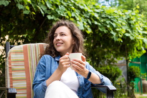 Portrait of dreamy young woman enjoying drinking coffee in the morning sitting on chair on home terrace on green garden background.Happy morning routine,day planning,enjoying life,to be in the moment
