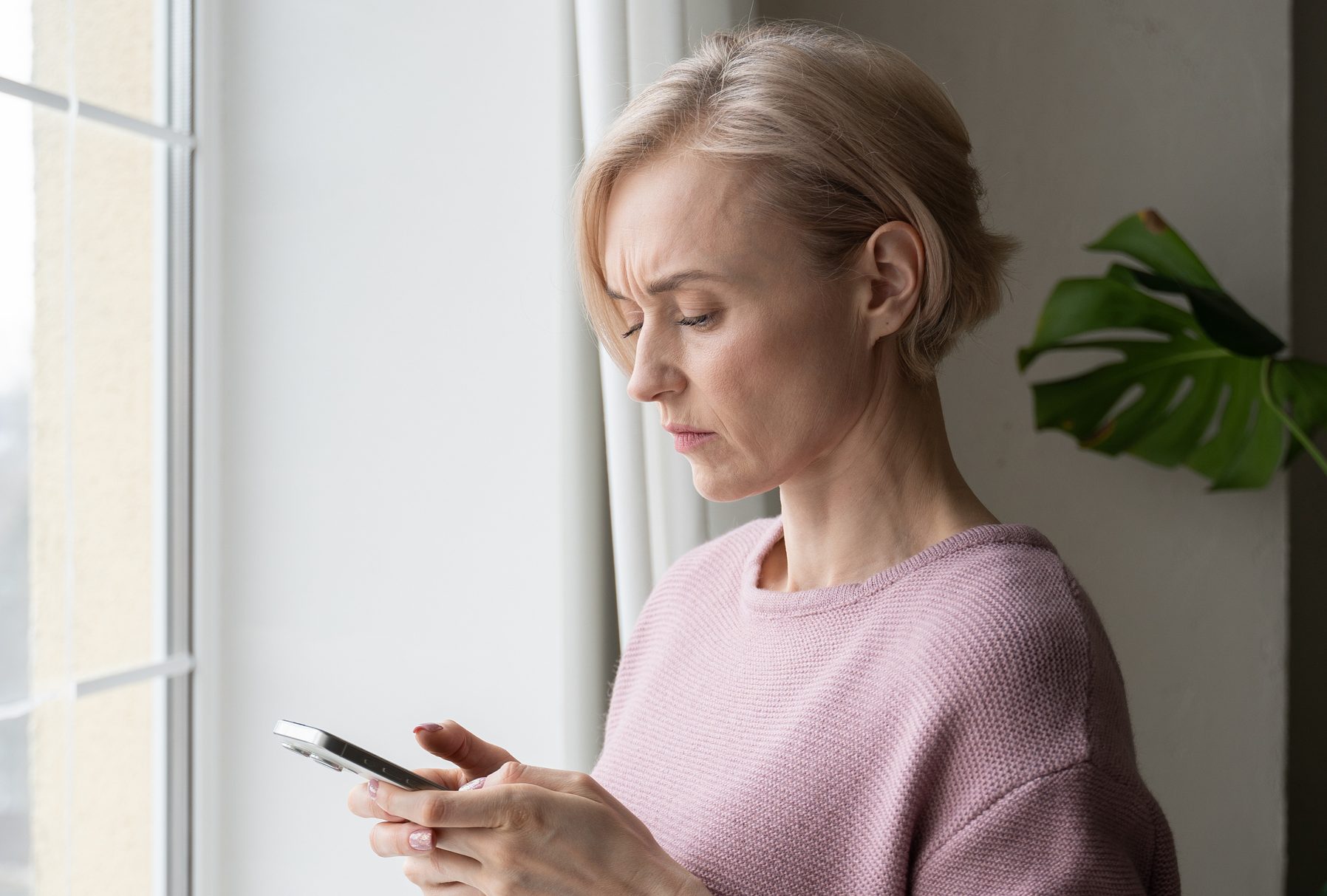 Worried Elderly Woman Checking Smartphone Beside Window on Overcast Day
