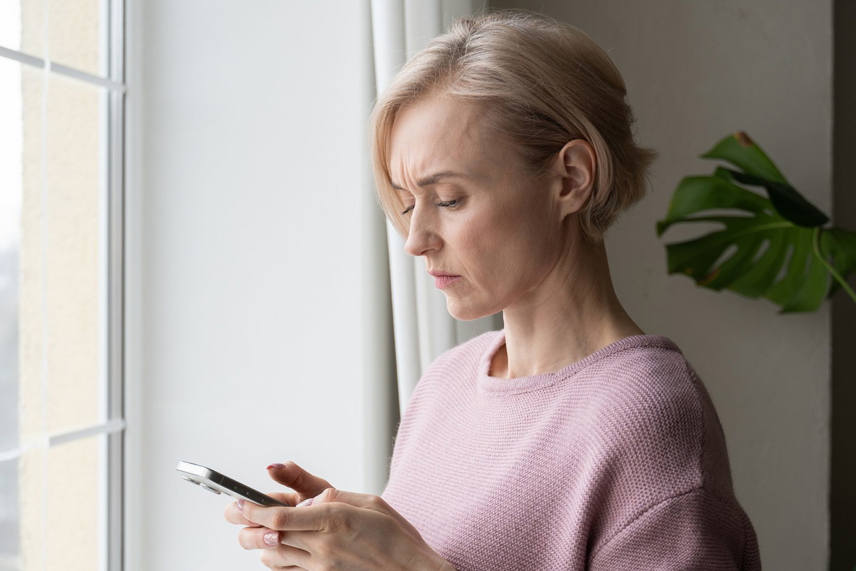 Worried Elderly Woman Checking Smartphone Beside Window on Overcast Day
