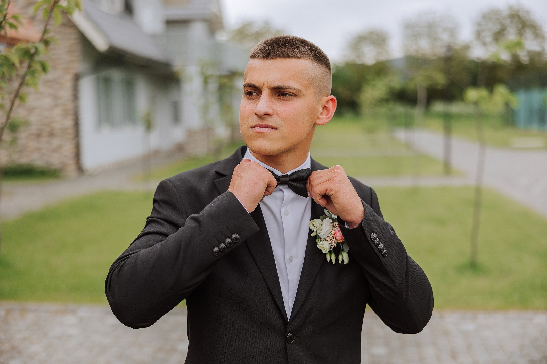 The groom in a black suit adjusts his bow tie, poses against the background of a green tree. Wedding portrait.