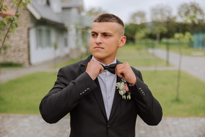 The groom in a black suit adjusts his bow tie, poses against the background of a green tree. Wedding portrait.