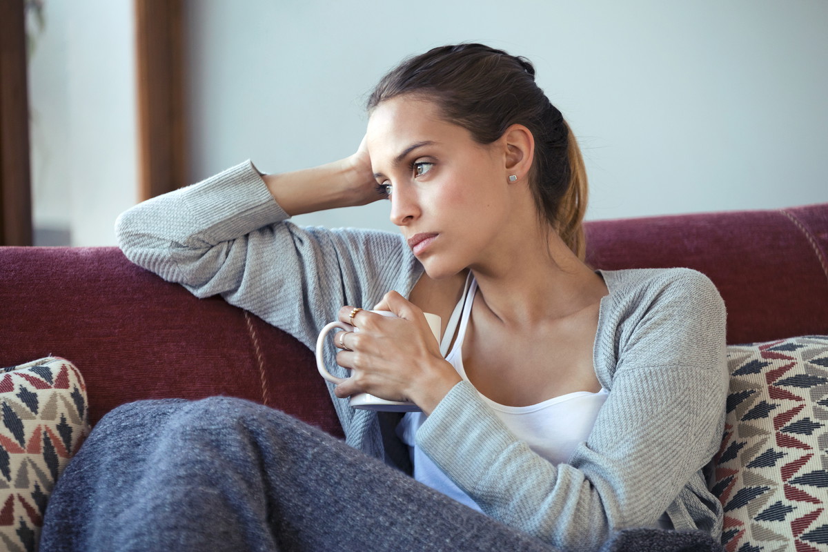 Depressed young woman thinking about her problems while drinking coffee on sofa at home.