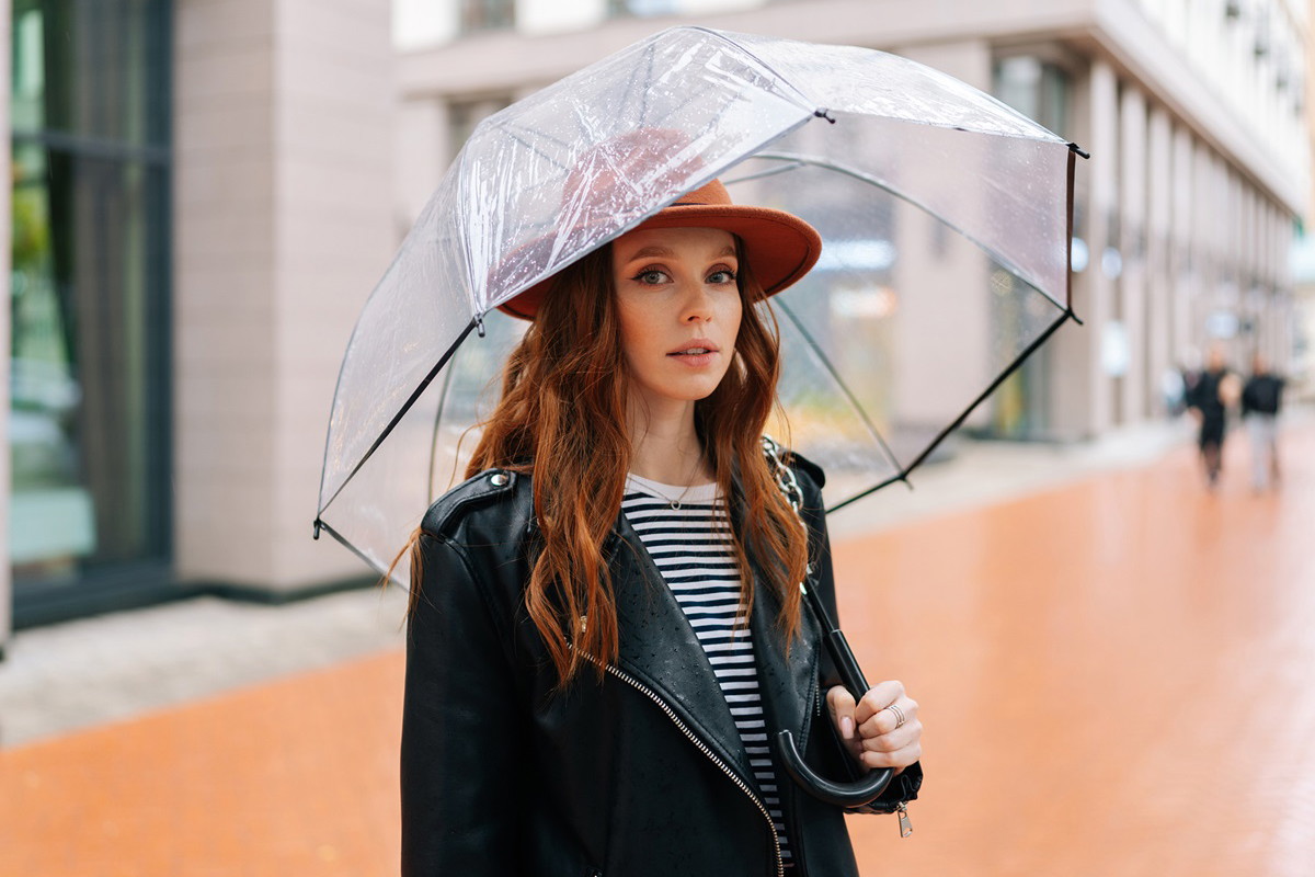 młoda dziewczyna o rudych włosach w kapeluszu i z parasolem stoi na ulicy zdezorientowana