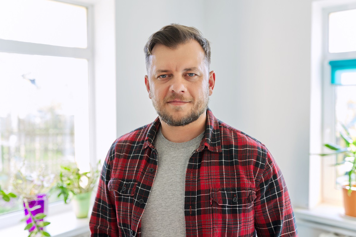 mężczyzna po 40 w kraciastej koszuli stoi w kuchni zamyślony z lekkim uśmiechem