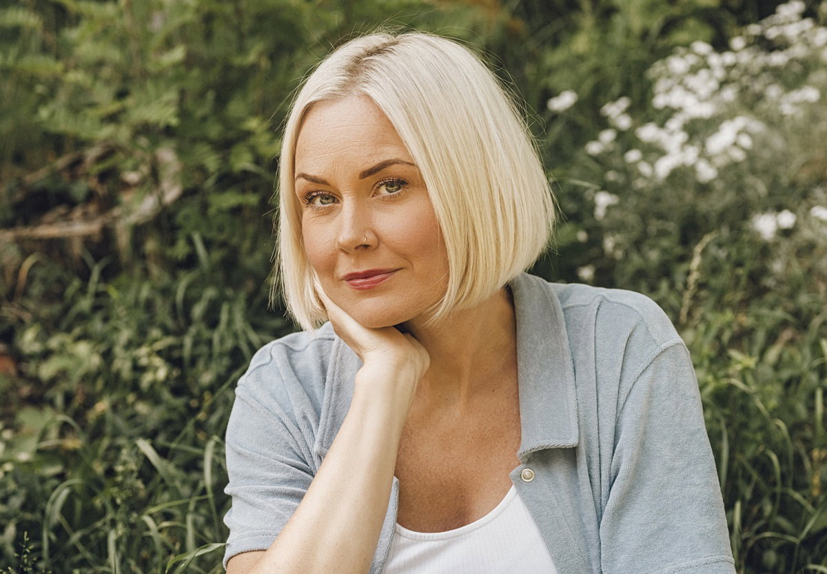 kobieta  w wieku około 40 lat blondynka siedzi na tle zieleni i patrzy podejrzliwie