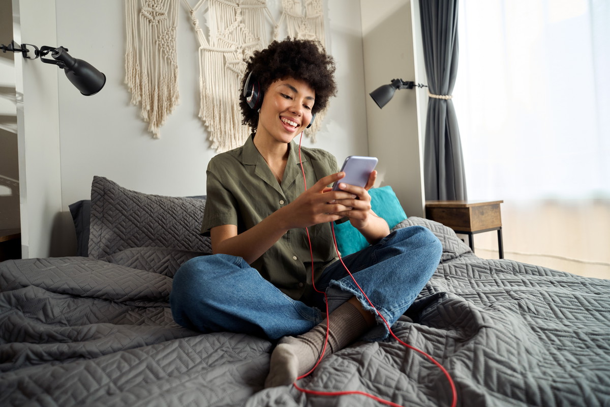Young happy funky African woman wearing headphones using phone at home.