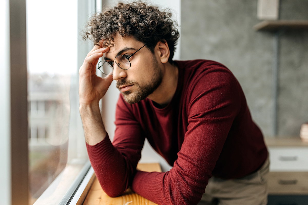 Portrait of depressed man contemplating his thoughts while standing next to the window in the apartment
