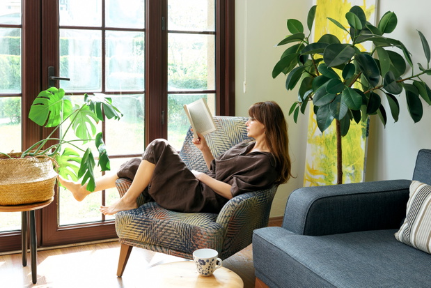 A woman relaxes in an armchair by a large window, engrossed in reading a book, surrounded by lush green houseplants.