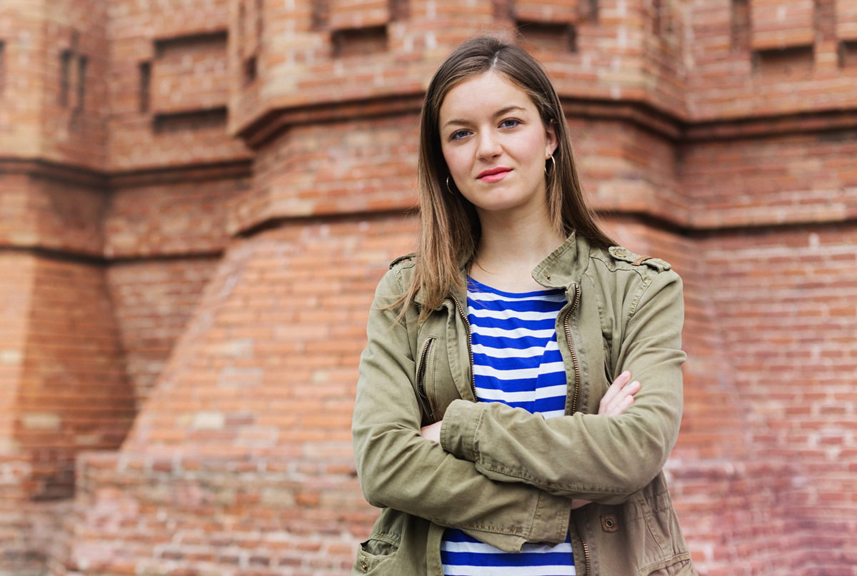 młoda dziewczyna w wieku 18 plus w zielonej kurtce stoi na tle murów poważna i zadziorna