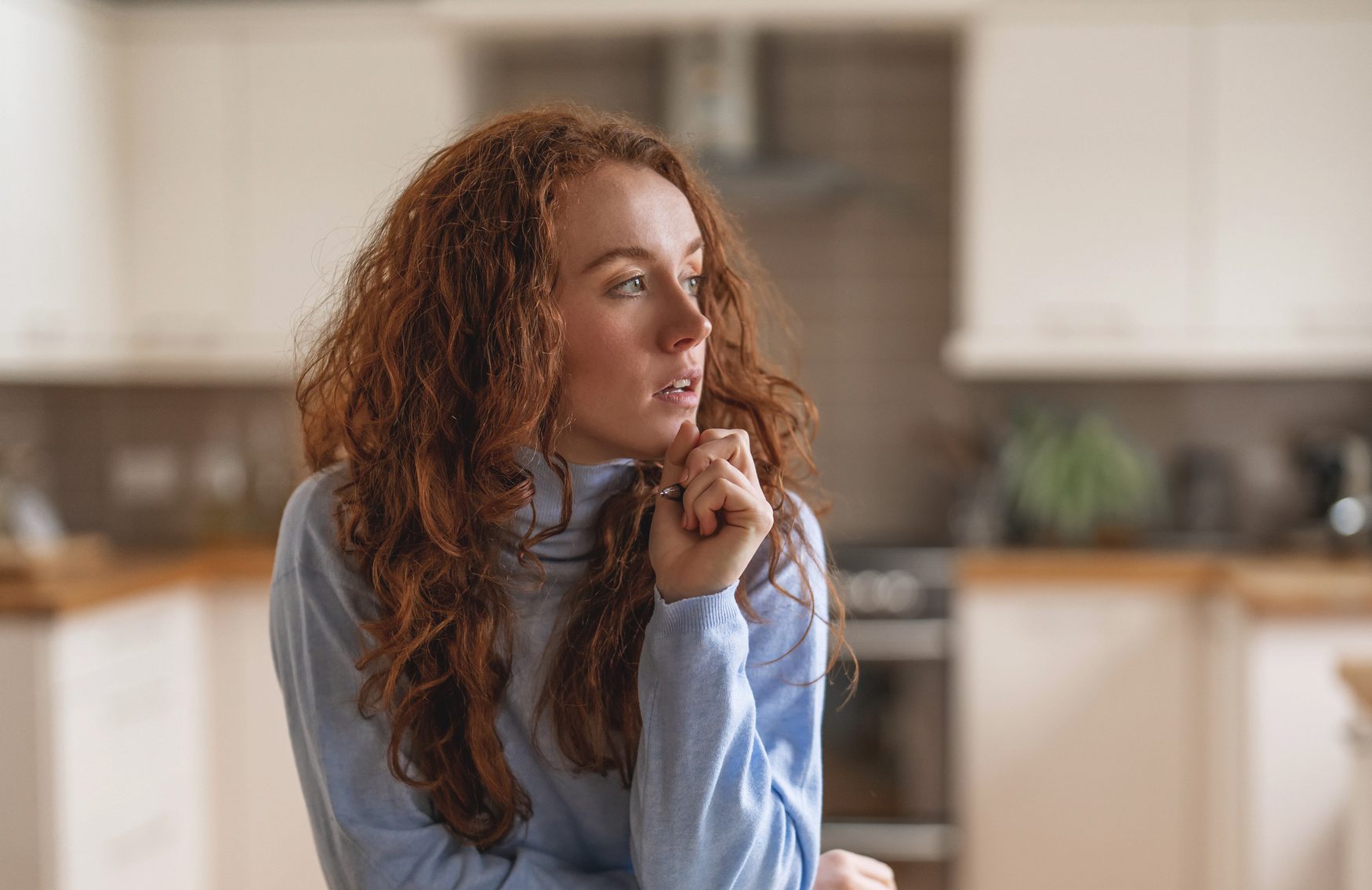 Woman with red hair thoughtfully gazes while seated at a wooden kitchen table in a modern home during midday