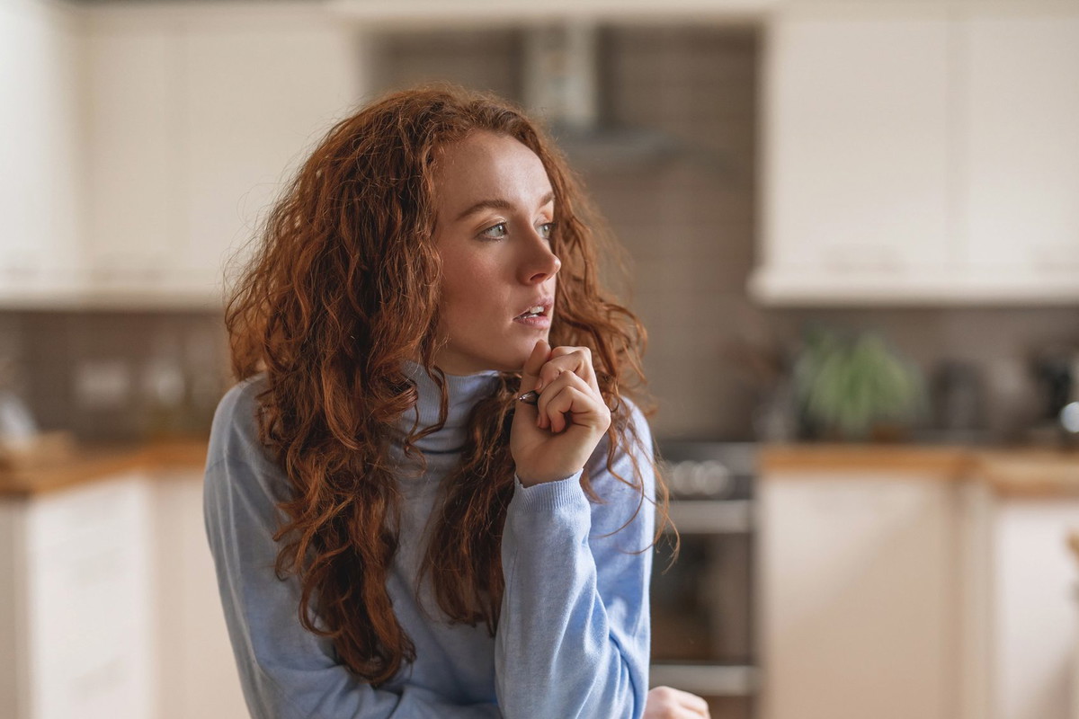 Woman with red hair thoughtfully gazes while seated at a wooden kitchen table in a modern home during midday