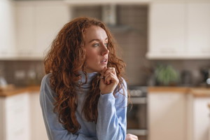 Woman with red hair thoughtfully gazes while seated at a wooden kitchen table in a modern home during midday